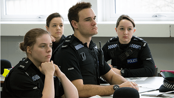 Four policing students in uniform sat at desks in a classroom looking towards a whiteboard